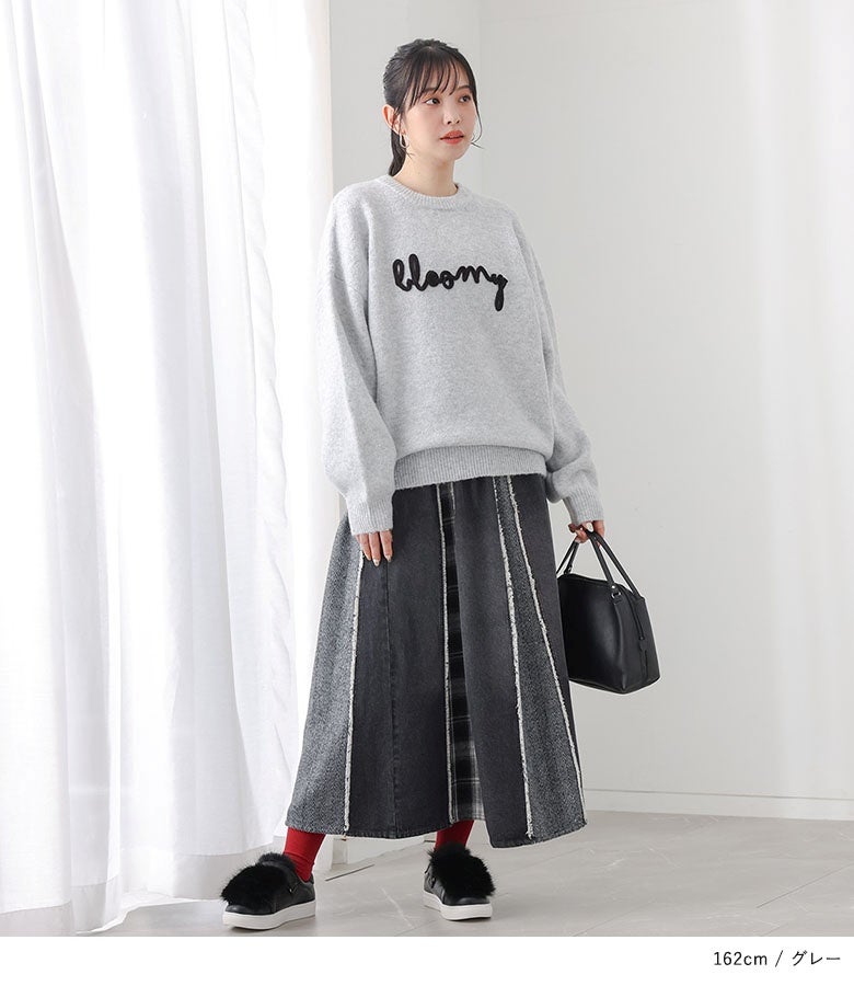 グレーのコード刺繍ロゴニットに、切り替えデザインのロングスカートを合わせたコーディネート。足元は黒のフェイクファーシューズと赤ソックスで華やかさをプラスしています。