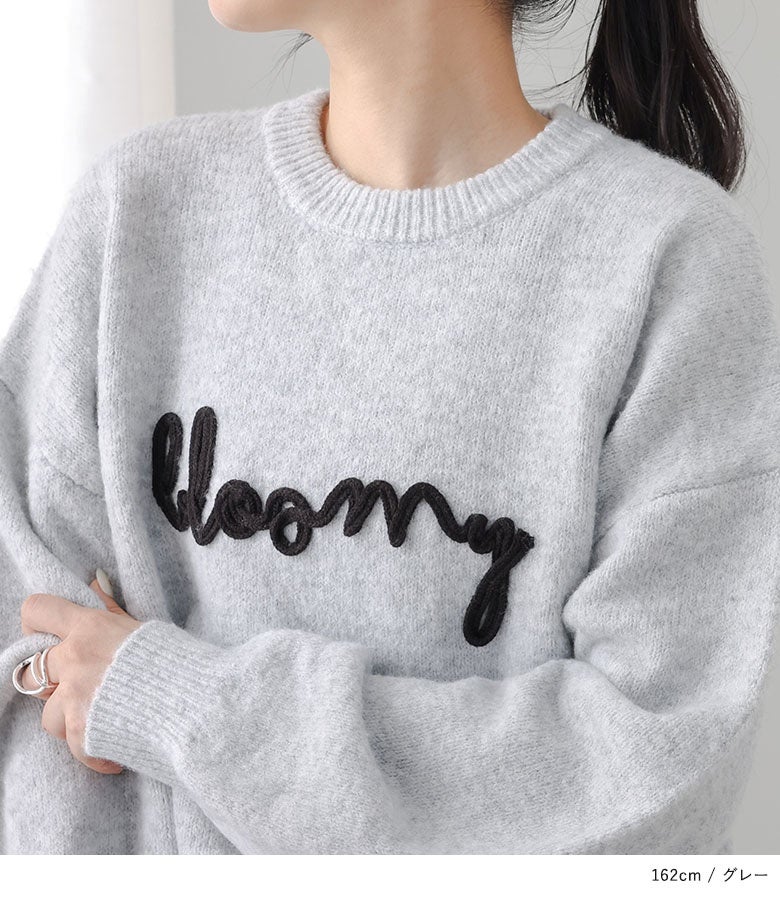 ライトグレーのニットトップスに配された、ブラックの立体ロゴ刺繍の近接ショット。ふんわり柔らかな質感と立体感のある刺繍が上質な雰囲気を漂わせます。